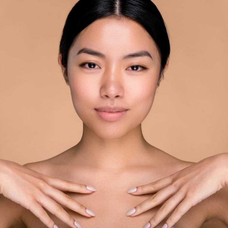 Beauty, care skin concept. Beautiful chinese brunette young woman, with perfect skin and naked shoulders, posing with hands near collarbone at isolated beige background, look at camera, smile pleasant