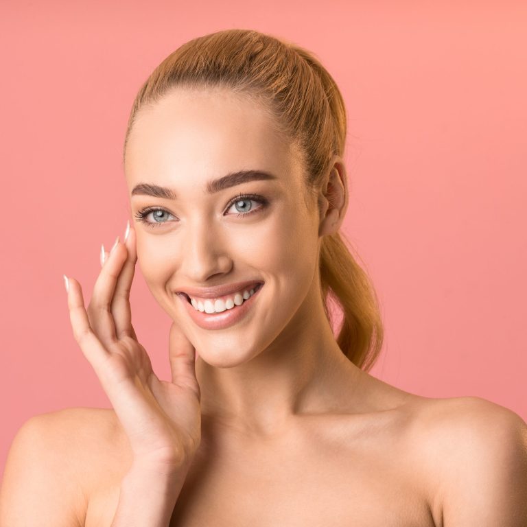 Daily Skincare. Smiling Lady Applying Facial Cream Looking Aside Posing In Studio Over Pink Background. Copy Space